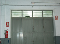 Puerta de entrada al taller con la señalización de seguridad de prohibido fumar a un lado y de extintor al otro. Además se indica la salida de emergencia y se observa la luz en la parte superior de la puerta.