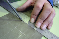 Imagen ampliable del trazado del plano a la pieza. La pieza está colocada sobre una superficie plana, y una mano sujeta una regla con la que se va trazando el dibujo sobre la cara plana de la pieza.