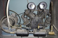 Foto de un compresor de émbolo doble. Se ve el equipo con dos cilindros que salen del cuerpo principal con dos manómetros.