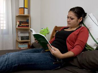 Vemos a una chica recostada en su cama con un libro abierto que está leyendo. Va vestida con pantalón vaquero, camiseta negra y chaqueta roja. Tiene el pelo oscuro y recogido.