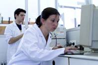 Apreciamos dos jóvenes, chico y chica. La joven está en primer plano. Tiene el pelo recogido, viste bata blanca y está manipulando un equipo de análisis. Están en una zona de trabajo identificable como laboratorio.