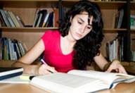 Vemos a una joven de cabello negro y largo. Viste una camiseta rosa fuerte. Está sentada a la mesa de lo que parece una biblioteca o una zona de estudio. Está leyendo un libro grande y tiene un bolígrafo en su mano derecha. 
