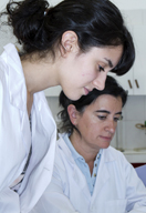 La imagen muestra a una joven junto a una compañera de mayor edad, ambas con bata blanca en lo que parece un laboratorio, observando algo que no se visualiza pero sobre lo que fijan ambas su mirada.