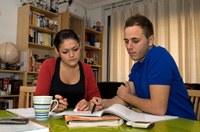La imagen muestra a dos jóvenes sentados, hojeando unos libros. Están en el domicilio de uno de ellos, tomando una taza de café a la vez que examinan la documentación.