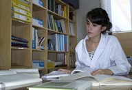 Vemos a una mujer joven que lleva bata blanca trabajando con varios libros abiertos sobre la mesa.