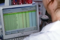 La imagen muestra una mujer sentada ante una pantalla de oredenadro con una tabla de datos. Solo se parecia parte del perfil izquierdo y por detrás, lleva una  bata blanca.