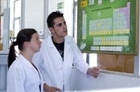 Apreciamos dos jóvenes, chico y chica, ambos con bata blanca observando la tabla periódica de los elementos químicos. Están en una zona de trabajo identificable como laboratorio.