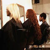 Imagen de tres chicas sentadas frente a un espejo en la peluquería, con una capa negra puesta sobre los hombros.  La primera tiene el cabello rubio, la segunda rojizo y largo y la tercera muy corto y oscuro.