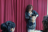 Imagen de una chica de cabello rizado, delante de un telón violeta, explicando sobre un maniquí alguna técnica, mientras la observan varias alumnas.