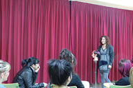 Imagen de una chica sobre un escenario con telón morado,  detrás de un maniquí dando clase a un grupo de mujeres.