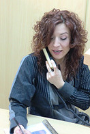  Imagen de una chica de cabellos rizados, sentada hablando por teléfono y tomando nota.  Tiene una chaqueta azul con rayas.