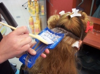 Imagen de una clienta con cabello largo, a la que una profesional le está realizando mechas con una paleta ancha azul. Con la mano derecha sujeta la paletina y aplica el decolorante.