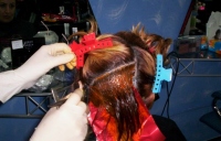  Imagen de un cabello visto por detrás, al que se le está realizando una técnica en diagonal. El cabello se separa con papel de estaño rojo, mientras unas manos con guantes sujetan el peine que separa el pelo. Tiene una grapa azul y otra roja.
