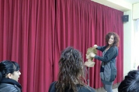 Imagen correspondiente al caso práctico. Se ve a una mujer con cabello rizado, vestida de gris, que explica a un grupo de alumnos, sobre un escenario con telón morado y con un maniquí entre las manos.