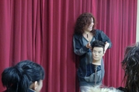 Imagen correspondiente al caso práctico, en la que se ve a una mujer explicando ante un grupo de alumnos. Está vestida de gris, tiene el cabello rizado y sujeta un maniquí. Detrás hay un telón morado.