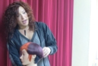 Imagen correspondiente al caso práctico. Se ve una chica con cabello rizado, vestida de gris, que está hablando. Delante de ella hay un maniquí con el cabello de varios colores. Detrás un telón morado.