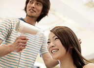 Se aprecia un profesional de la peluquería, secándole el cabello a su clienta que, está con una sonrisa en la boca.