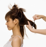 Se ve una mujer de perfil, lleva el cabello sujeto en varias particiones. La partición de la nuca está suelta y unas manos manipulan el cabello.